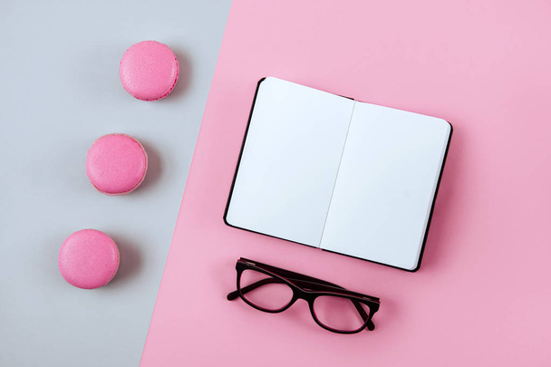 Macaron, clean notebook, eyeglasses on pink table from above. Female working desk. Flat lay style. - Zdjęcie, obraz