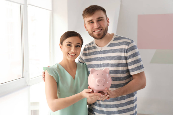 Casal feliz segurando banco porquinho dentro de casa. Conceito de poupança monetária
 - Foto, Imagem