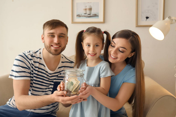 Happy little girl with her parents holding glass jar with dollars indoors. Money savings concept - Фото, изображение