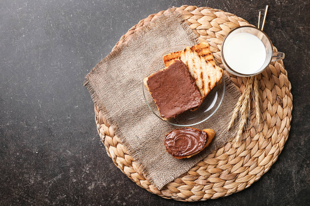 Поджаренный хлеб с шоколадной пастой и чашкой молока на плетеном коврике
 - Фото, изображение