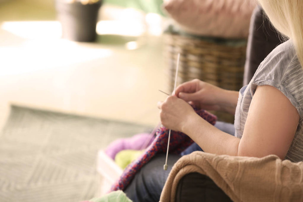 Mujer madura sentada en el sofá y tejiendo suéter caliente en el interior
 - Foto, Imagen