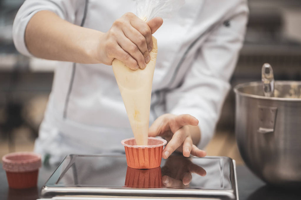 Nahaufnahme der Hände des Küchenchefs mit Zuckertüte, die Sahne auf Cupcakes drückt - Foto, Bild