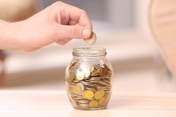 Man putting coins into glass jar on table. Savings concept - Фото, изображение