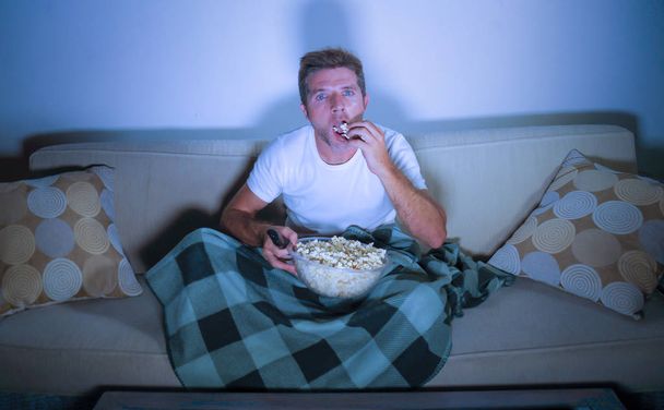 lifestyle portrait of young attractive relaxed man watching movie on television eating popcorn sitting late night at living room sofa couch enjoying fully concentrated and interested - Foto, afbeelding
