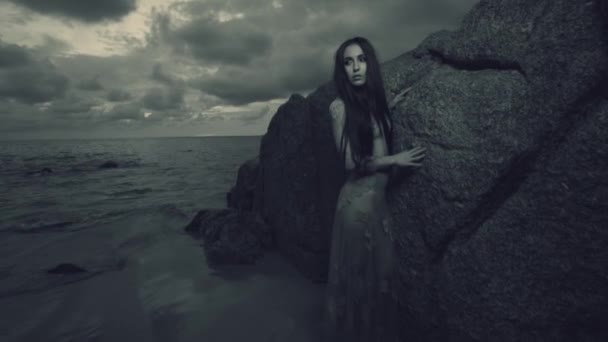 Tajemná krása na pláži při západu slunce / krásná tajemná žena v dlouhých šatech na písečné pláži poblíž skály nad moře a západ slunce oblohou pozadí - černé a bílé video v pomalém pohybu - Záběry, video