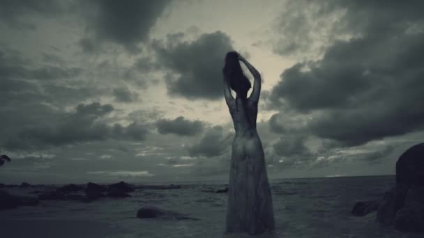 Beleza misteriosa na praia durante o pôr do sol / Visão traseira da bela mulher misteriosa em vestido longo na praia de areia perto de rochas sobre o mar e céu nublado fundo preto e branco vídeo em câmera lenta
 - Filmagem, Vídeo
