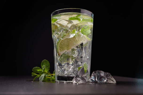 Glass of iced Mojito cocktail and ingredients,  lime, ice and mint leaves on black background, cold summer drink  - Φωτογραφία, εικόνα