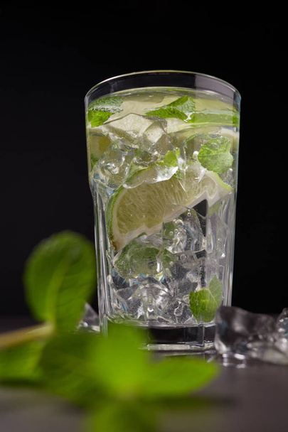Glass of iced Mojito cocktail and ingredients,  lime, ice and mint leaves on black background, cold summer drink  - Foto, Imagem