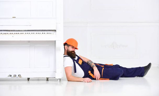Loader lean on piano instrument. Man with beard, worker in overalls and helmet fall asleep tired, white background. Courier fall asleep while moving furniture, relocation. Tired worker concept - Photo, Image