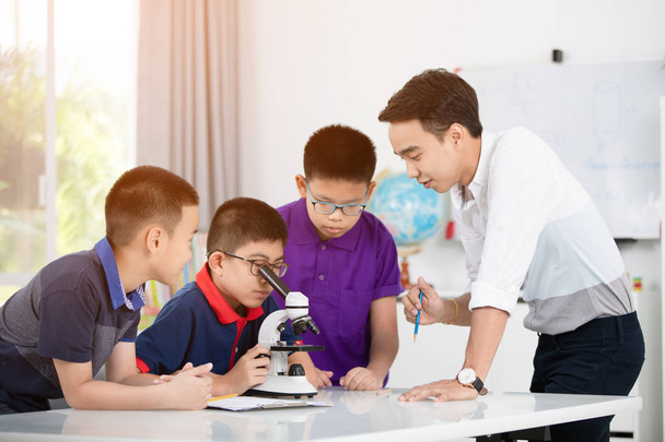 Веселый мужчина-учитель объясняет что-то группе молодых учеников в классе. Азиатский мальчик рассматривает подготовку под микроскопом
 - Фото, изображение