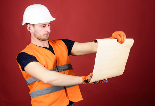 Man in helmet, hard hat holds building plan, controls works, red background. Engineer, architect, builder on strict face holds old blueprint in hands, supervises construction site. Foreman concept - Photo, image