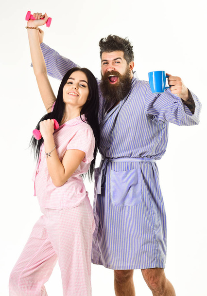 Couple, family on sleepy faces, full of energy. Couple in love in pajama, bathrobe stand isolated on white background. Morning routine concept. Girl with dumbbell, man with coffee cup - 写真・画像