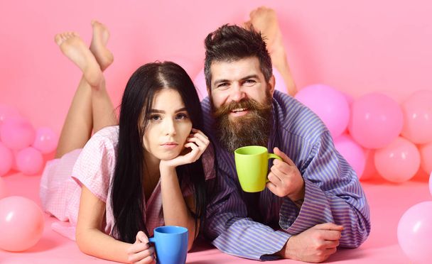 Couple relax in morning with coffee. Man and woman on smiling faces lay, pink background. Weekend morning concept. Couple in love drink coffee in bed. Man and woman in domestic clothes, pajamas - Photo, image
