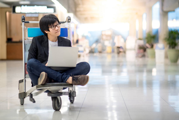 Молодой азиат, работающий на тележке аэропорта со своим ноутбуком в ожидании стыковочного рейса, внештатного образа жизни и цифровых концепций кочевников
 - Фото, изображение