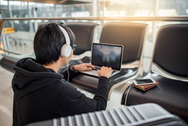 Küçük Asya adam Uluslararası Havaalanı terminal, seyahat yaşam biçimi uçuş bağlanmak için beklerken bankta dizüstü bilgisayar kullanan kulaklık ile - Fotoğraf, Görsel
