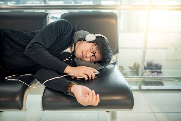 Young Asian man with eyeglasses and headphones sleeping on bench while waiting for connecting flight in the international airport terminal, travel lifestyle - Fotó, kép
