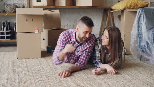 Een gelukkig paar is liggend op de vloer in het nieuwe huis, man geeft sleutels aan zijn vriendin en kuste haar, ze praat en lacht. Huisvesting en relatie concept. - Video