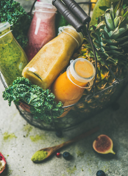 Colorful smoothies in bottles with fresh tropical fruit and greens in basket over grey concrete background - Fotó, kép