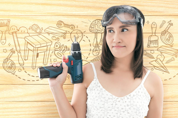 Asian woman dressed in white top holding a cordless electric drill on wood background - Photo, Image