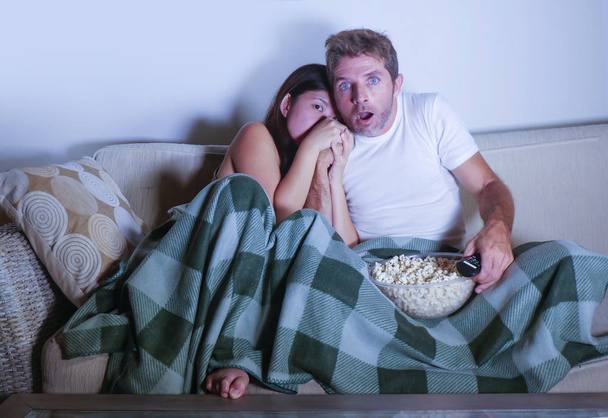 young mixed race attractive couple with Asian Korean woman and white man enjoying together watching television horror movie scared girlfriend in panic covering eyes on sofa couch eating popcorn  - Фото, изображение
