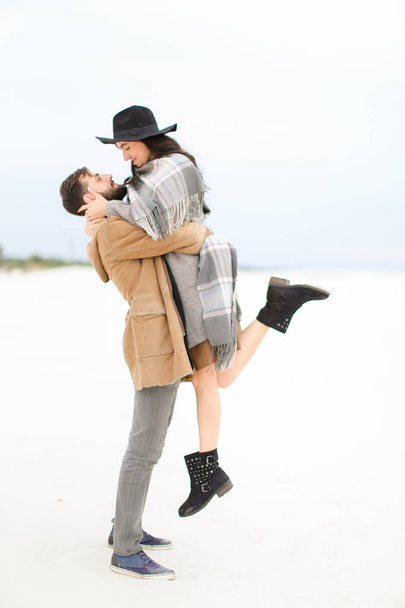 Man with beard picking up young woman wearing hat, coat and grey scarf on white snow background. - Photo, Image