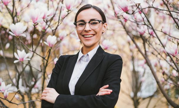 Young caucasian businesswoman in suit and glasses smiling with crossed arms among blooming magnolia trees - Fotó, kép
