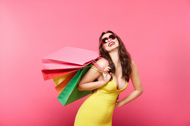 mladá žena nosí sluneční brýle drží barevné tašky proti růžové pozadí, nakupování a slevy koncept - Fotografie, Obrázek