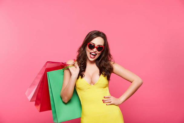 young woman wearing sunglasses holding colorful shopping bags against pink background, shopping and discount concept - Foto, afbeelding
