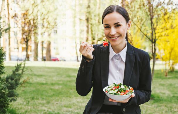 Young caucasian brunette businesswoman in suit eating salad outdoors. Concept of healthy eating during work. - Photo, Image