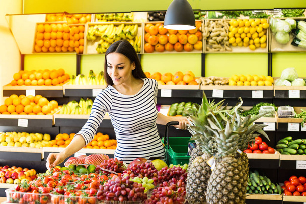 Smiling caucasian brunette woman with basket trying to choose vegetables in grocery - Valokuva, kuva