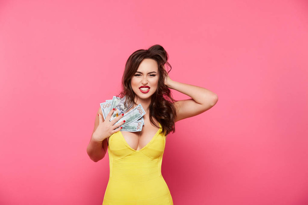 привлекательная привлекательная женщина в желтом платье позирует на розовом фоне держа наличные деньги, торговый концепт
 - Фото, изображение