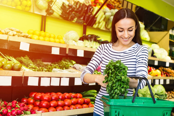 Smiling caucasian brunette woman with basket trying to choose vegetables in grocery - Photo, image