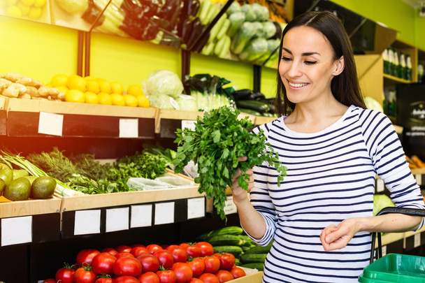 Smiling caucasian brunette woman with basket trying to choose vegetables in grocery - Fotografie, Obrázek