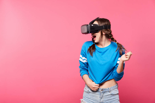 young woman with with virtual reality glasses posing at pink background, 3D, future concept - Фото, изображение