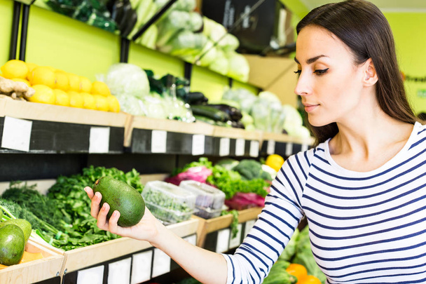 Smiling caucasian brunette woman with basket trying to choose vegetables in grocery - Foto, Imagen