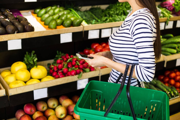 Smiling caucasian brunette woman with basket trying to choose vegetables in grocery - Photo, Image