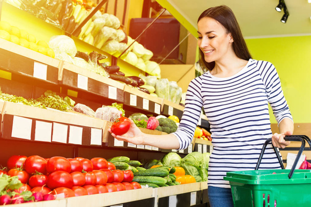 Smiling caucasian brunette woman with basket trying to choose vegetables in grocery - Foto, afbeelding