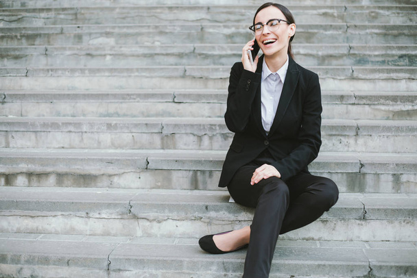 Young cheerful caucasian brunette businesswoman with phone sitting on stairs outdoors - Foto, imagen