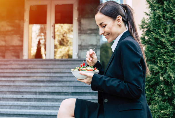Young caucasian brunette businesswoman in suit eating salad outdoors. Concept of healthy eating during work. - Foto, Bild