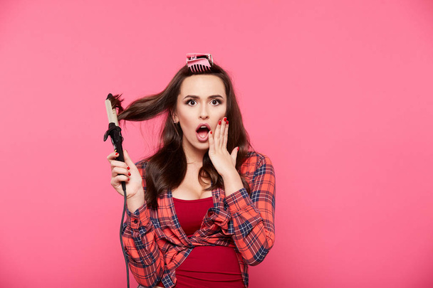 gorgeous young woman with curly hair posing at pink background and doing her hair straighter - Photo, Image