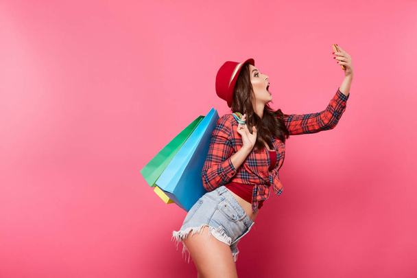 junge glückliche Frau mit bunten Einkaufstaschen mit Handy vor rosa Hintergrund, Einkaufs- und Rabattkonzept - Foto, Bild