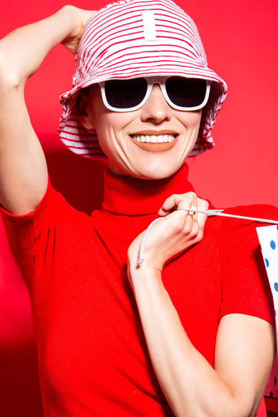 Parlak kırmızı arka planda alışveriş torbaları olan, şapkalı ve güneş gözlüklü mutlu bir kadın. - Fotoğraf, Görsel