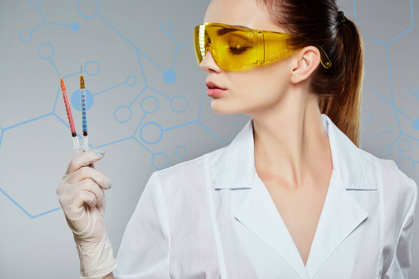 Женщина-врач в белой медицинской форме, желтые защитные очки и держа два шприца на сером фоне студии
 - Фото, изображение