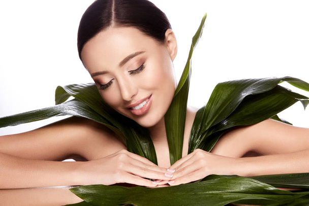 красивая и естественная молодая женщина позирует с зелеными листьями на сером фоне, концепция ухода за кожей, увлажненная кожа
 - Фото, изображение