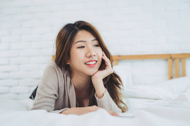 Portrait beautiful young Asian woman on bed at home in the morning. Cheerful Asian woman wearing a comfortable sweater and smiling on her bed. Relaxing room. lifestyle asia woman at home concept. - Fotó, kép