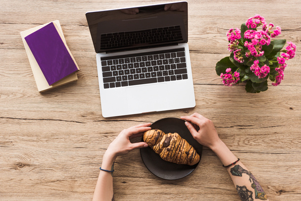 Schnappschuss einer Frau am Arbeitsplatz mit Laptop, Büchern und Croissant auf dem Teller - Foto, Bild