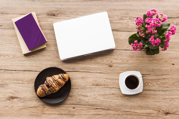 flat lay with laptop, books, cup of coffee, croissant on plate and kalanchoe plant on wooden tabletop - Photo, Image