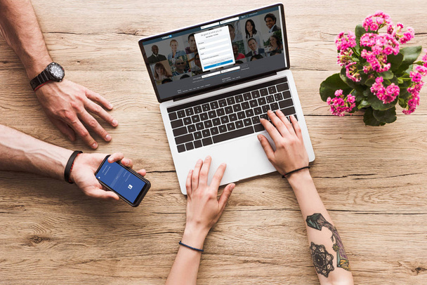 cropped shot of man with smartphone with facebook logo in hand and woman at tabletop with laptop with linkedin website and kalanchoe flower - Foto, Imagem