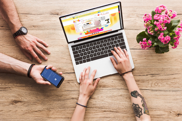 cropped shot of man with smartphone with facebook logo in hand and woman at tabletop with laptop with aliexpress website and kalanchoe flower - Foto, Imagen