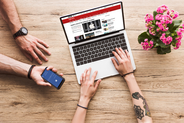 cropped shot of man with smartphone with facebook logo in hand and woman at tabletop with laptop with bbc website and kalanchoe flower - Fotografie, Obrázek